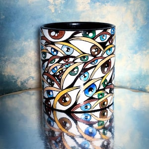 May include: A black ceramic mug with a white background and a pattern of colorful eyes. The eyes are brown, blue, and green.