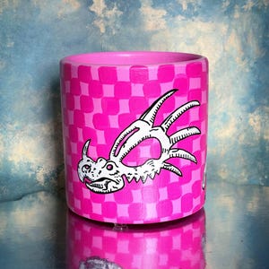 May include: A cylindrical, pink ceramic pot with a checkered pattern and a white and black dragon illustration. The pot's design is reflected on the shiny surface below. The background is a cloudy blue.