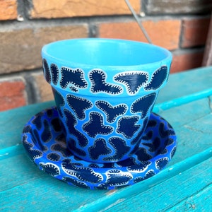 Peut inclure: Un pot en céramique bleu avec un motif abstrait noir et argent. Le pot a une soucoupe assortie.