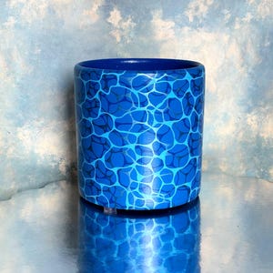 May include: A cylindrical container with a vibrant blue water-like pattern. The design features interconnected shapes in varying shades of blue. The interior and rim are a solid, darker blue. The container is reflected on a shiny surface.