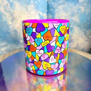 Peut inclure: Une tasse en céramique rose avec un motif coloré de cœurs portant différents messages. Les messages incluent "Love", "Only You", "True Love", "Be Mine", "Hug Me", "You & Me", "Kiss Me", et "XOXO".