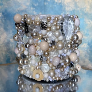 May include: A cylindrical decorative container covered in a mosaic of pearls, beads, and crystals in shades of white, cream, and silver. The surface is highly textured with varying sizes and shapes of embellishments. A large, clear crystal is prominent on the side.