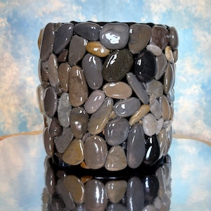 May include: A cylindrical container decorated with smooth, polished river stones in shades of gray, brown, and black. The stones are arranged closely together, creating a textured surface. The container sits on a reflective surface.