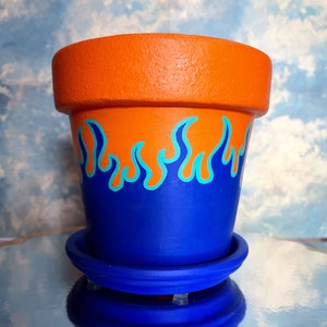 Peut inclure: Pot de fleurs en terre cuite peint en orange et bleu avec une soucoupe bleue. Le pot est peint avec un motif de flammes en bleu et turquoise.