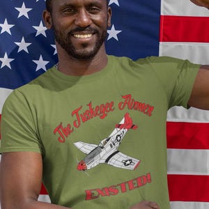 May include: A green t-shirt with a white and red airplane graphic and the text "The Tuskegee Airmen Existed!" printed on it. The shirt is being worn by a person standing in front of an American flag.