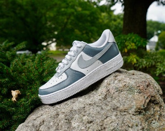 May include: A white and gray low-top sneaker with a gray swoosh logo. The shoe is sitting on a gray rock.