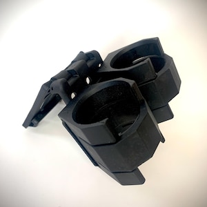 Könnte beinhalten: Eine mattschwarze Kunststoff-Klemmhalterung mit zwei kreisförmigen Öffnungen und einem Klappmechanismus. Das Gerät hat ein strukturiertes, geometrisches Design mit sichtbaren Gelenken und einem Verriegelungselement.