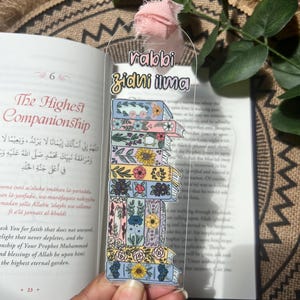 May include: Clear acrylic bookmark featuring a stack of illustrated books with floral designs. The bookmark has the text "rabbi zidni ilma" at the top and a pink ribbon. The bookmark is held over an open book.