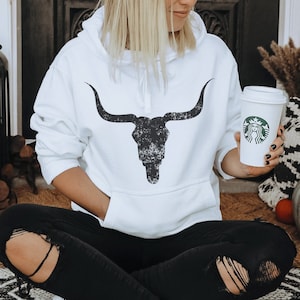 Pu&ograve; includere: Felpa bianca con una grafica di teschio di toro nero invecchiato. La felpa ha un cappuccio con coulisse e una tasca a marsupio.