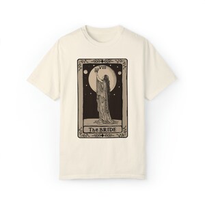 Op de afbeelding: Een beige t-shirt met een zwart-witte illustratie van een tarotkaart. De kaart toont een vrouw die een kelk vasthoudt en de tekst "VIII The BRIDE".