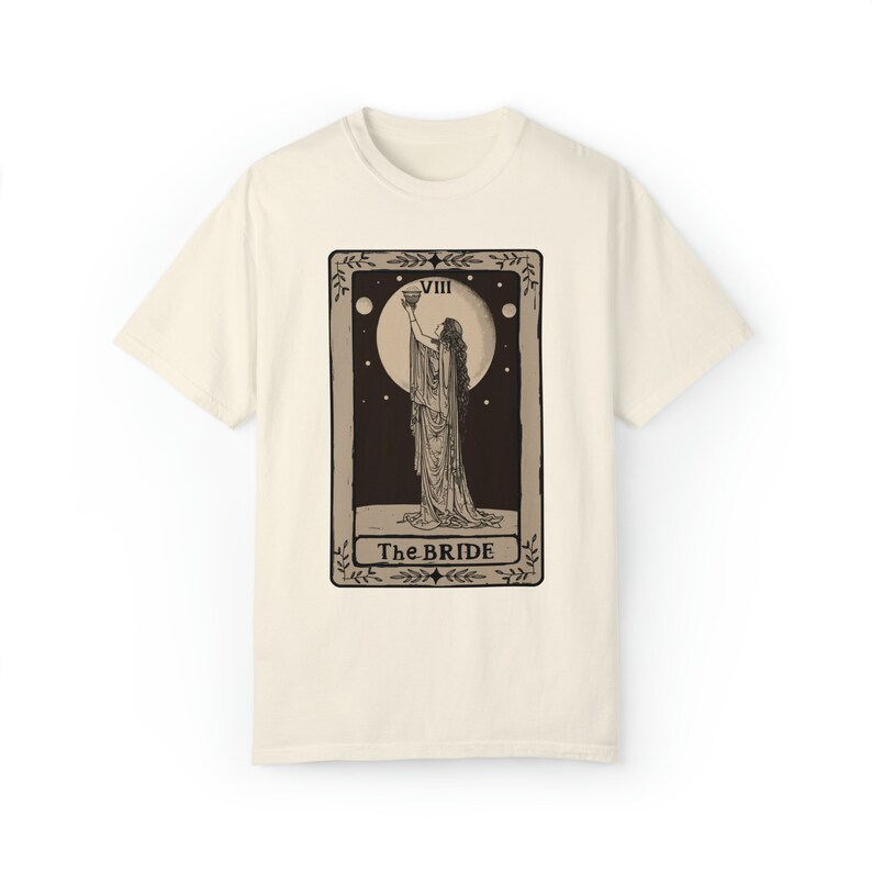 Op de afbeelding: Een beige t-shirt met een zwart-witte illustratie van een tarotkaart. De kaart toont een vrouw die een kelk vasthoudt en de tekst "VIII The BRIDE".