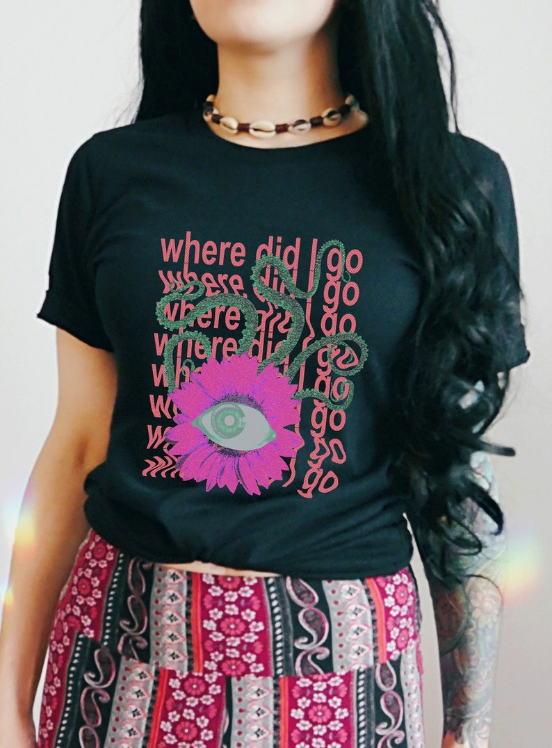 May include: Black t-shirt featuring a pink flower and eye graphic with the text "Where did I go" repeated in a pattern. The model is wearing a colourful patterned skirt.