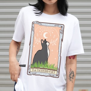 May include: White t-shirt with a graphic of a black and white cat with a crescent moon and stars. The text "The Tuxedo Cat" is printed below the cat.