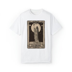 Op de afbeelding: Wit T-shirt met een bruin tarotkaartontwerp met een vrouw die een beker vasthoudt en de tekst "VIII The BRIDE".