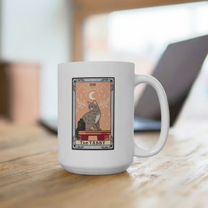Può includere: Tazza da caffè in ceramica bianca con un'illustrazione colorata di una carta dei tarocchi con un gatto. La carta presenta un gatto tabby grigio seduto su una piattaforma con una pianta. La carta è intitolata "The Tabby" e ha il numero romano "VIII" nell'angolo superiore.