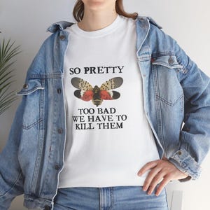 Può includere: T-shirt bianco con grafica di insetto e la scritta "SO PRETTY TOO BAD WE HAVE TO KILL THEM". La maglietta è abbinata a una giacca di jeans blu e jeans blu.