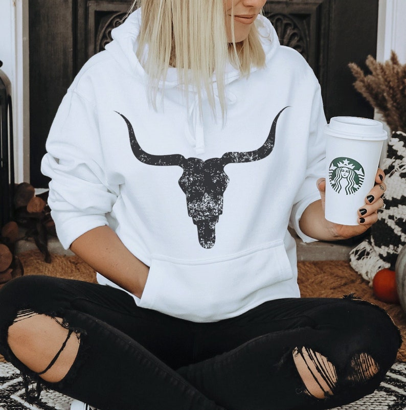 Pu&ograve; includere: Felpa bianca con una grafica di teschio di toro nero invecchiato. La felpa ha un cappuccio con coulisse e una tasca a marsupio.
