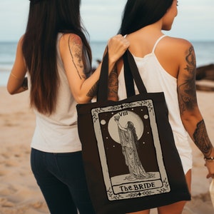 Può includere: Una borsa tote nera con un design di carta dei tarocchi che mostra una donna che tiene un calice sotto una luna piena. La carta recita "VIII La Sposa".