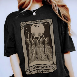 Op de afbeelding: Zwart T-shirt met een vintage tarotkaartontwerp met een volle maan en vier figuren die glazen heffen. De kaart luidt "XIV The BRIDESMAID".