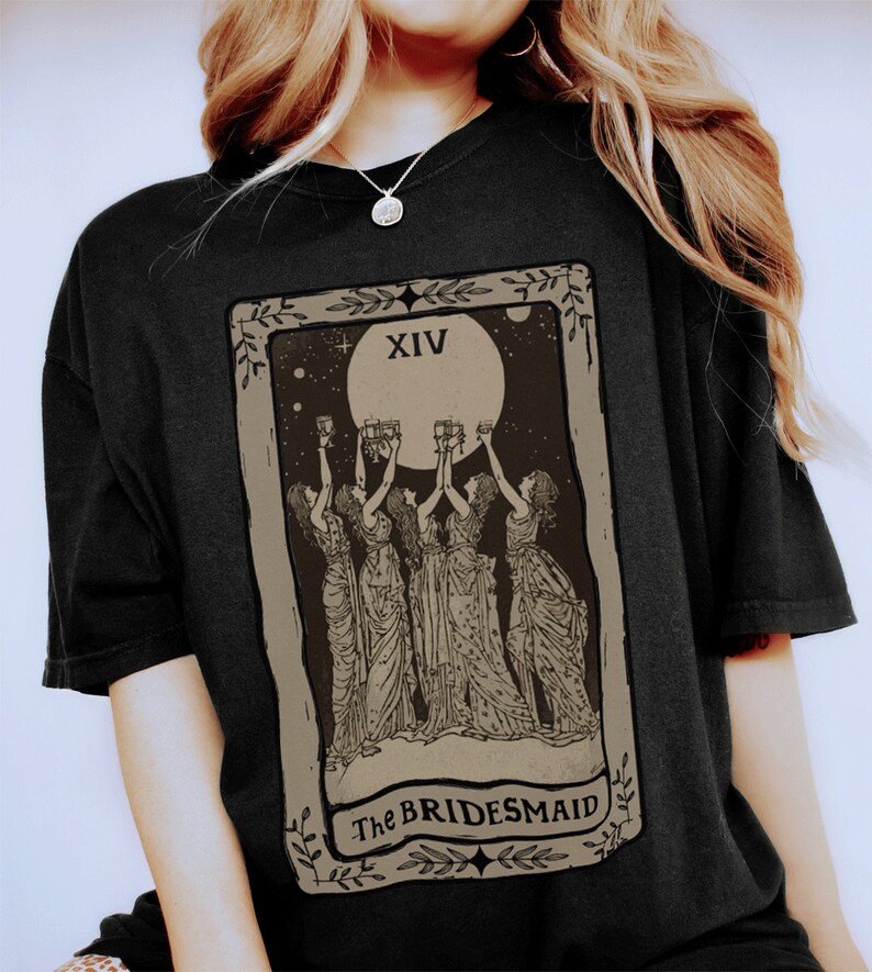 Op de afbeelding: Zwart T-shirt met een vintage tarotkaartontwerp met een volle maan en vier figuren die glazen heffen. De kaart luidt "XIV The BRIDESMAID".