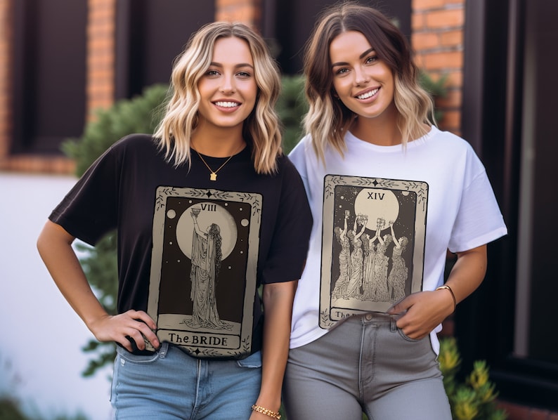 Op de afbeelding: Twee vrouwen dragen zwarte en witte t-shirts met tarotkaartontwerpen. Het zwarte shirt heeft de kaart "De Bruid" met nummer VIII en een vrouw die een kelk vasthoudt. Het witte shirt heeft de kaart "De Keizerin" met nummer XIV en vier vrouwen die kelken vasthouden.