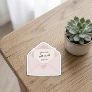 May include: A die-cut sticker shaped like a light pink envelope with a white border. Inside the envelope is a cream-colored note with the handwritten black text "You've got mail" and "xoxo" below it. A small pink heart is centered on the envelope flap.
