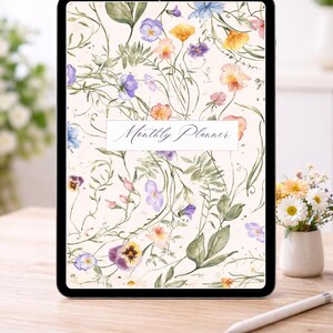 May include: A digital tablet displaying a floral design with the words "Monthly Planner." The design features watercolor flowers in shades of purple, yellow, and pink, with green leaves on a cream background. A stylus and a small vase of flowers are on the wooden table.