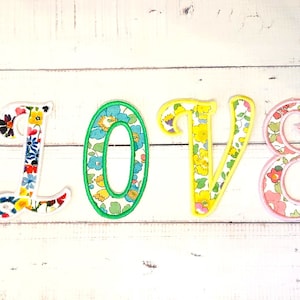 May include: Fabric letters spelling "LOVE" in a cursive style. Each letter is filled with a floral pattern in shades of blue, yellow, and pink, and outlined in matching colours. The letters are displayed on a white wooden surface.