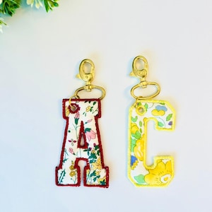 Könnte beinhalten: Zwei Stoff-Schlüsselanhänger in Form der Buchstaben A und C. Der Buchstabe A hat ein florales Muster auf weißem Grund mit einem dunkelroten gestickten Rand. Der Buchstabe C hat ein gelb-weißes Blumenmuster mit einem gelben gestickten Rand.