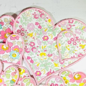May include: A collection of ten heart-shaped fabric patches in varying sizes, each featuring a white background with a colourful floral pattern in shades of pink, yellow, and green, finished with a stitched pink border around every edge.