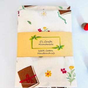May include: A white cotton handkerchief with a brown book, red flowers, and yellow flowers print. The handkerchief is folded in half and has a brown label that reads "C's Crafts #handmade 100% Cotton Handkerchiefs".