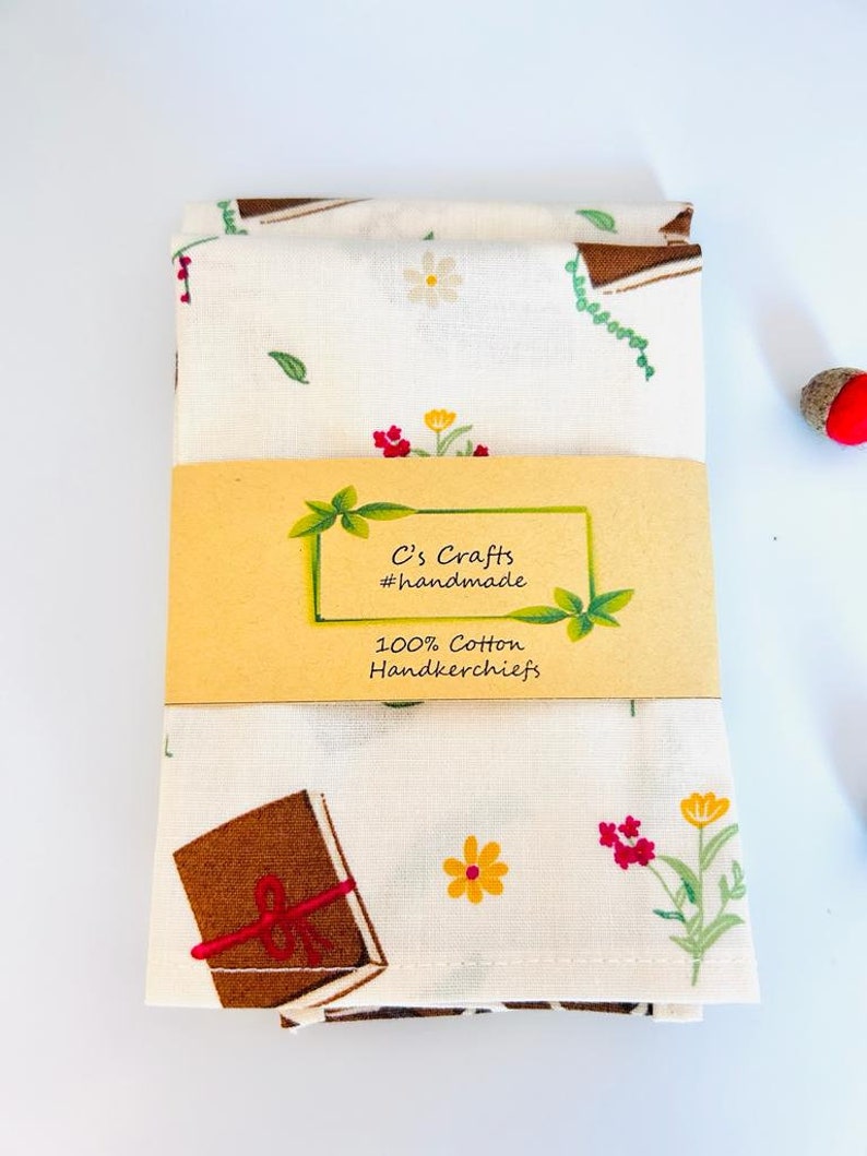 May include: A white cotton handkerchief with a brown book, red flowers, and yellow flowers print. The handkerchief is folded in half and has a brown label that reads "C's Crafts #handmade 100% Cotton Handkerchiefs".
