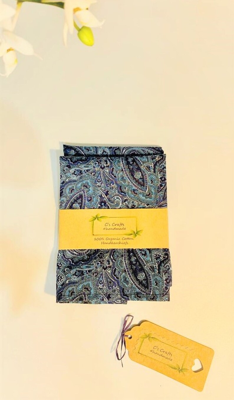 Op de afbeelding: Een set van twee blauwe paisley-gepatroneerde zakdoeken gemaakt van 100% biologisch katoen. De zakdoeken zijn gevouwen en op elkaar gestapeld. Een klein bruin label met de tekst "C's Crafts #handmade" is aan de zakdoeken bevestigd.