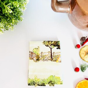 K&ouml;nnte beinhalten: Ein wei&szlig;es Geschirrtuch mit einer Aquarell-Illustration einer l&auml;ndlichen Szene mit einer Steinmauer, B&auml;umen und einem Holztor. Das Geschirrtuch ist gefaltet und liegt auf einer wei&szlig;en Oberfl&auml;che.