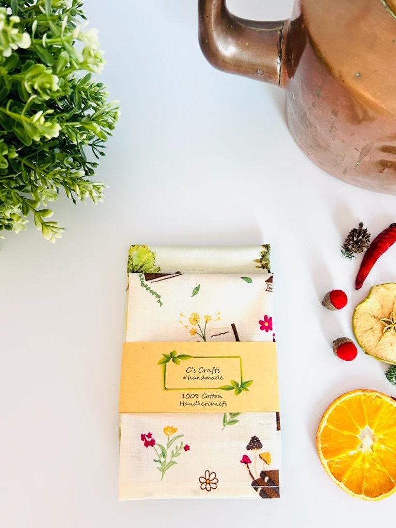 May include: A set of two white cotton handkerchiefs with a floral and mushroom print. The handkerchiefs are packaged with a yellow label that reads "C's Crafts #handmade 100% Cotton Handkerchiefs".