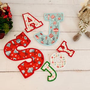 May include: Red and blue fabric letters spelling out "JOY" against a white background. The letters are decorated with a Christmas-themed pattern of baubles and stars.