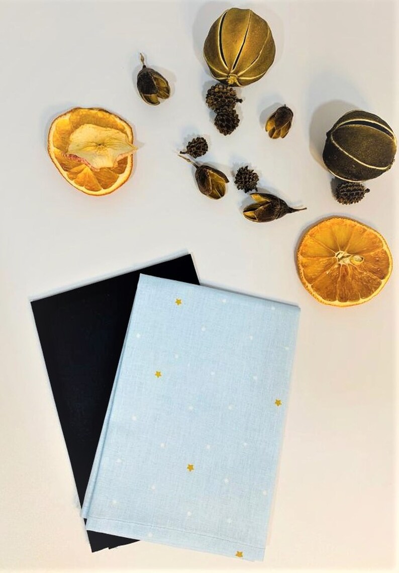 May include: A blue tea towel with white polka dots and gold stars. The tea towel is folded in half and is sitting on top of a black piece of fabric. There are dried orange slices and other decorative items on a white surface.
