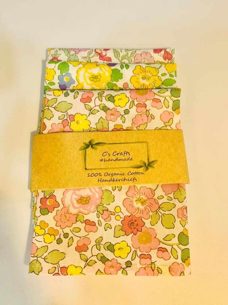 May include: A set of three floral handkerchiefs made from 100% organic cotton. The handkerchiefs are packaged with a brown paper label that reads "C's Crafts #handmade 100% Organic Cotton Handkerchiefs".