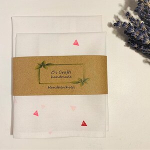 May include: Two white handkerchiefs with a pink and red triangle pattern. The handkerchiefs are folded and stacked on top of each other. A brown label with the text "C's Crafts handmade Handkerchiefs" is attached to the handkerchiefs.