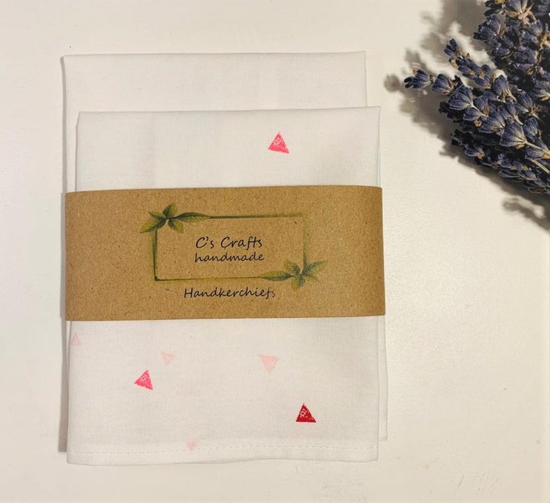 May include: Two white handkerchiefs with a pink and red triangle pattern. The handkerchiefs are folded and stacked on top of each other. A brown label with the text "C's Crafts handmade Handkerchiefs" is attached to the handkerchiefs.