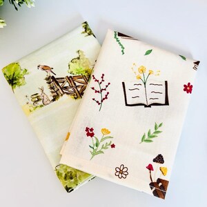 May include: Two fabric squares with different patterns. One square has a light green background with a watercolor illustration of a bird, rabbit, and a fence. The other square has a cream background with a book, flowers, and mushrooms.