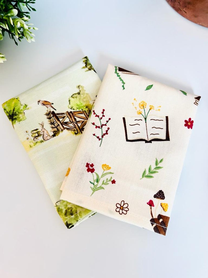 May include: Two fabric squares with different patterns. One square has a light green background with a watercolor illustration of a bird, rabbit, and a fence. The other square has a cream background with a book, flowers, and mushrooms.