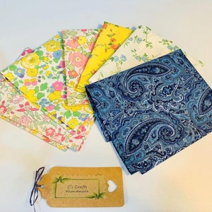Op de afbeelding: Een set van acht stoffen vierkanten, waaronder een blauwe paisley print en zeven bloemenprints in tinten roze, geel en wit. De stoffen vierkanten zijn in een waaier vorm gerangschikt. De tekst "C's Crafts #handmade" is zichtbaar op een bruin label.