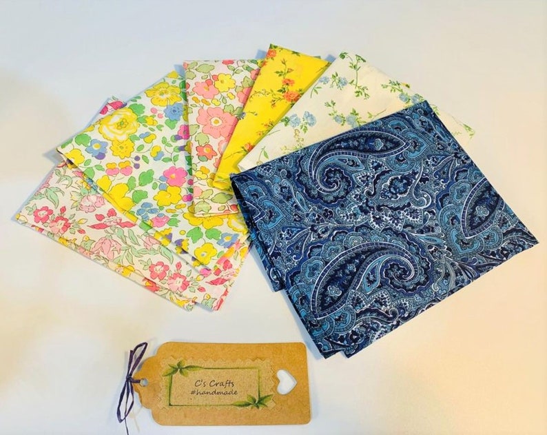 Op de afbeelding: Een set van acht stoffen vierkanten, waaronder een blauwe paisley print en zeven bloemenprints in tinten roze, geel en wit. De stoffen vierkanten zijn in een waaier vorm gerangschikt. De tekst "C's Crafts #handmade" is zichtbaar op een bruin label.