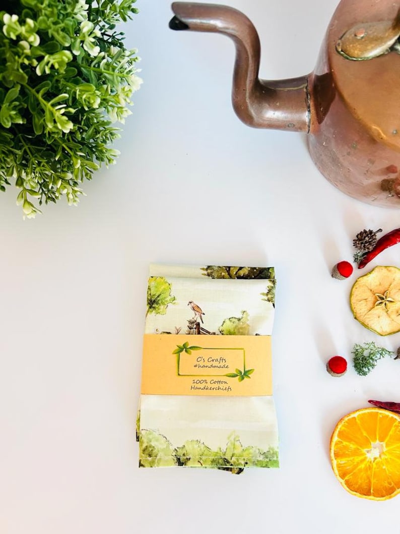 K&ouml;nnte beinhalten: Zwei gr&uuml;ne und wei&szlig;e Baumwolltaschent&uuml;cher mit einem Vogel- und Baumdesign. Die Taschent&uuml;cher sind mit einem gelben Etikett verpackt, auf dem "C's Crafts #handmade 100% Cotton Handkerchiefs" steht.