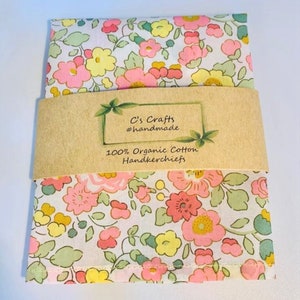 May include: A white handkerchief with a pink, yellow, and green floral pattern. The handkerchief is packaged with a brown paper label that reads "C's Crafts #handmade 100% Organic Cotton Handkerchiefs".