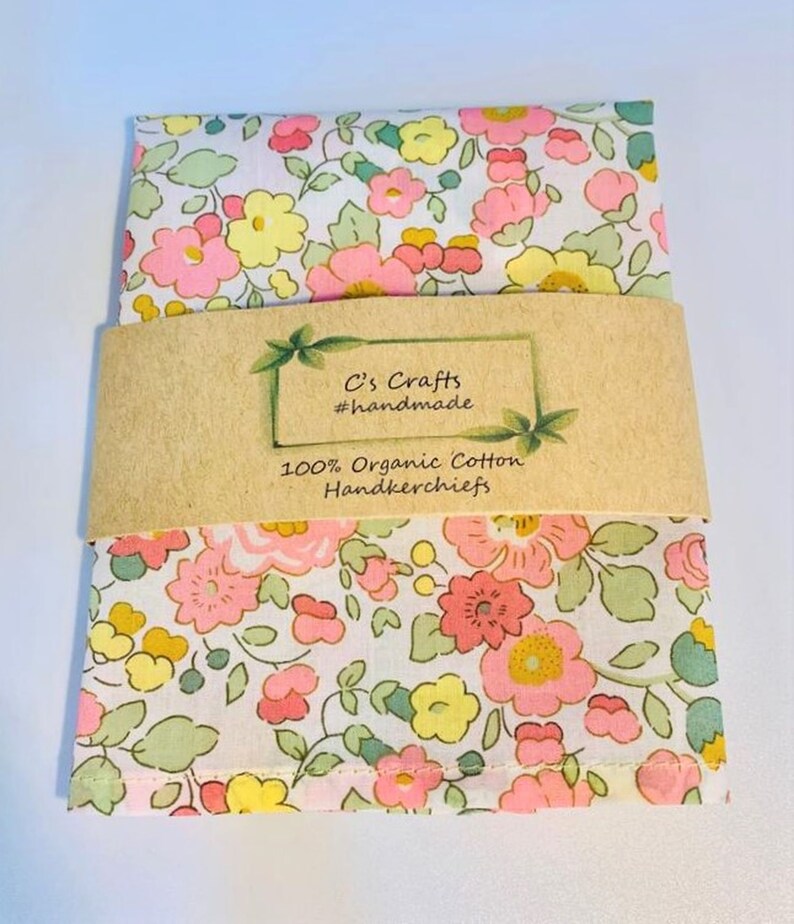 May include: A white handkerchief with a pink, yellow, and green floral pattern. The handkerchief is packaged with a brown paper label that reads "C's Crafts #handmade 100% Organic Cotton Handkerchiefs".