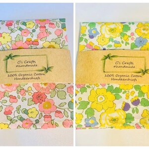 May include: Two floral handkerchiefs with a white background and colorful flowers. The handkerchiefs are packaged with a brown paper label that reads "C's Crafts #handmade 100% Organic Cotton Handkerchiefs".