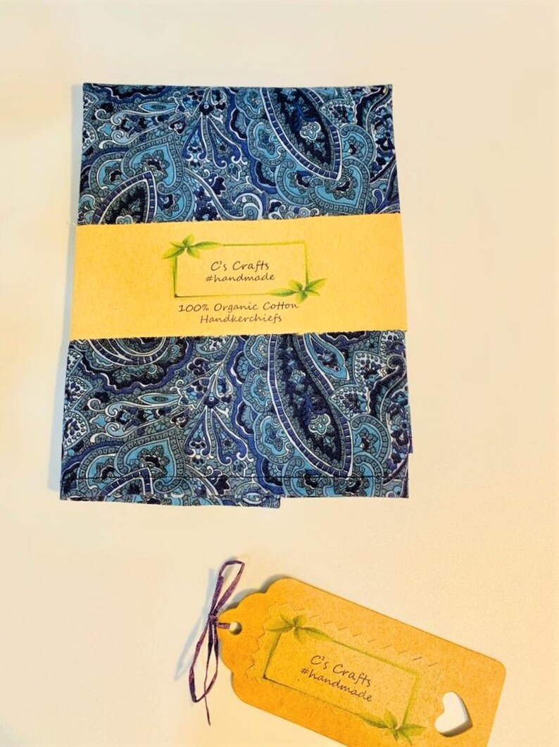 Op de afbeelding: Een blauwe paisley-gepatroneerd zakdoek gemaakt van 100% biologisch katoen. De zakdoek is gevouwen en verpakt met een bruin papieren label waarop "C's Crafts #handmade" staat.