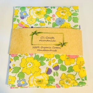 May include: A white cotton handkerchief with a yellow, pink, and blue floral pattern. The handkerchief is folded and has a brown paper label that reads "C's Crafts #handmade 100% Organic Cotton Handkerchiefs".